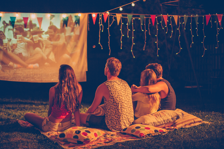 Young friends having movie night party. Laying down on blanket in front of movie improvised screen. Backyard decorated with festive string lights. Night time. View from behind.